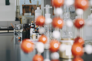 En kemi labb med mätglas, bägare, och mörka flaskor i bakgrunden, framför ett suddigt molekylmodell-set med röda och vita kulor.